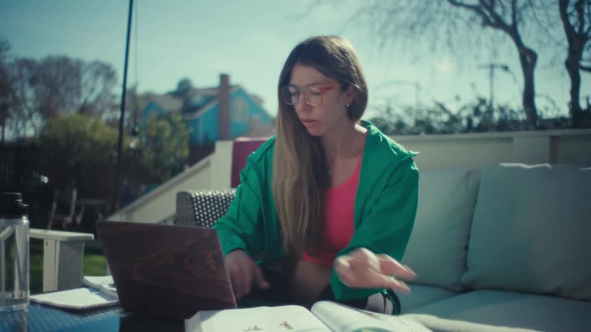 A woman wearing glasses and a green jacket sits on an outdoor patio flipping through a textbook and typing on a laptop computer while studying in the bright sunlight
