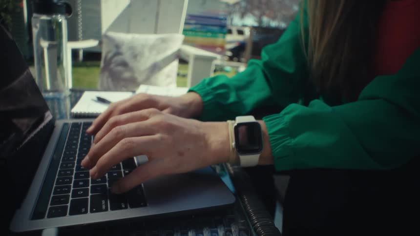 A woman wearing a green jacket and a white smartwatch types on a laptop computer at a glass table before closing the lid in a bright outdoor setting