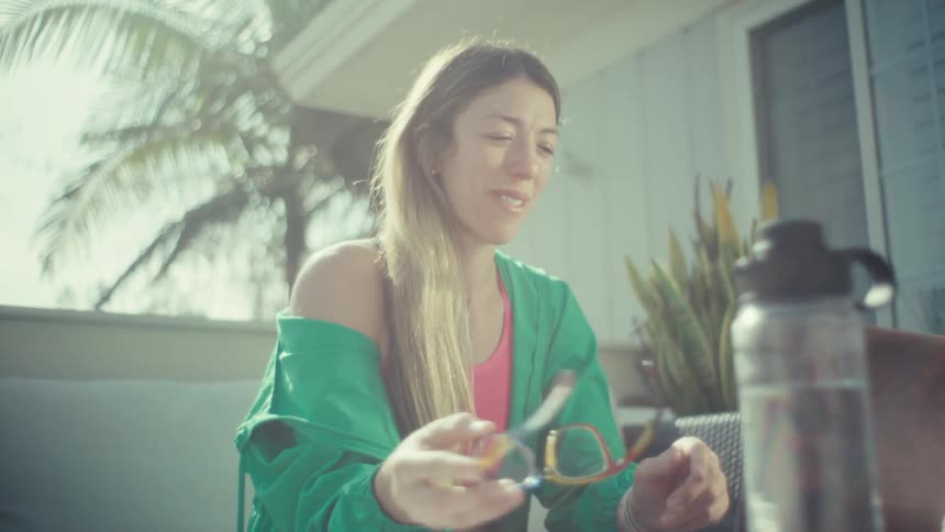 A woman wearing a green jacket and colorful glasses sits on an outdoor patio typing on a laptop computer before taking off her glasses and looking away with a pensive expression in the bright sunlight