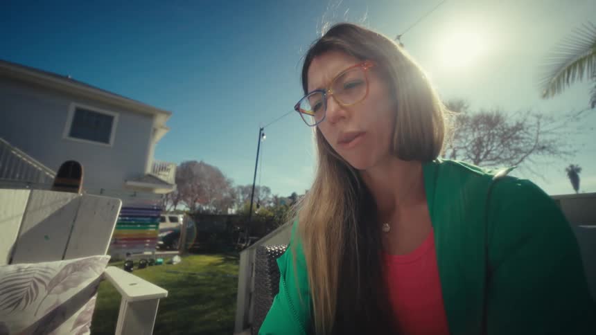 A young woman wearing glasses sits on a couch under the sun typing on her laptop