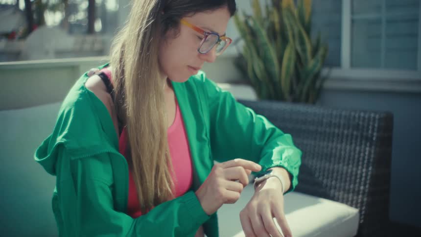 A woman wearing a green jacket and colorful glasses sits on an outdoor patio and looks at her white smartwatch while working at a laptop computer in the bright sunlight