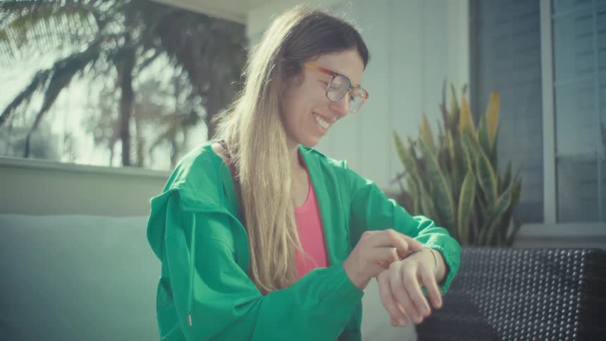 A pregnant woman wearing a green jacket and colorful glasses sits on an outdoor patio and types on a laptop computer before checking her smartwatch with a smile in the bright sunlight