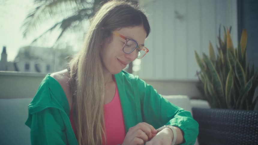 A woman wearing a green jacket and colorful glasses sits on an outdoor patio and looks at her smartwatch while working on a laptop computer in the bright sunlight