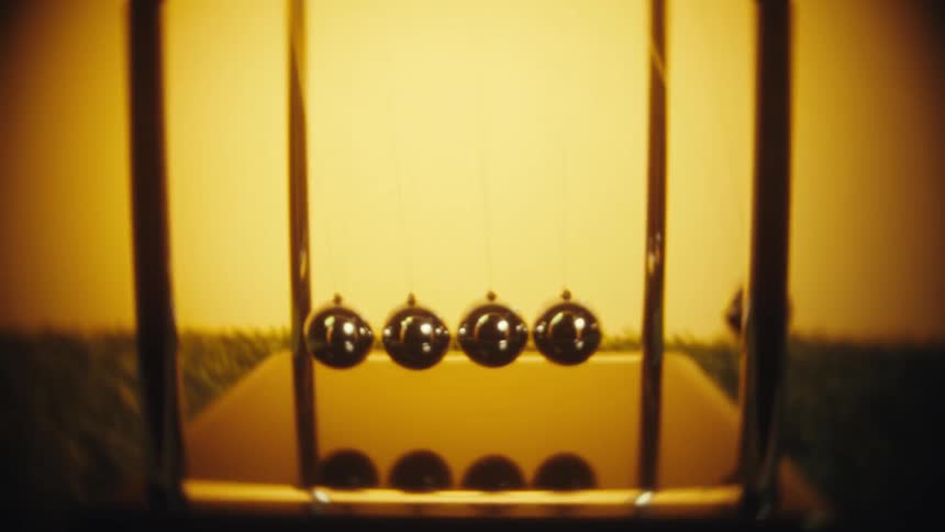 A person with a gold ring on their finger pulls back a silver ball on a newtons cradle and releases it to begin the rhythmic motion against a warm yellow background on a patch of green grass