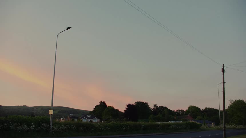 Cars passing on a road next to houses on a suburban area with hills on the background outdoors during dusk or dawn