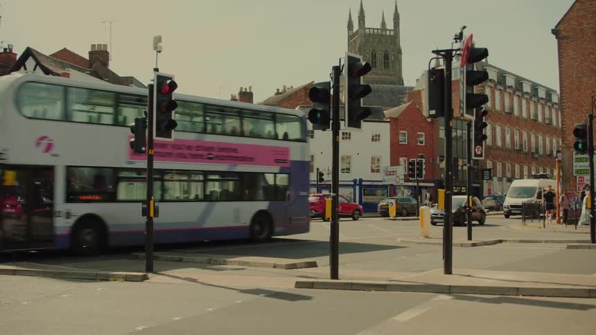 A blue doubledecker bus drives through a busy city intersection past traffic lights and red brick buildings while a tall stone church tower stands in the distant background under a clear sky during the day in an urban environment