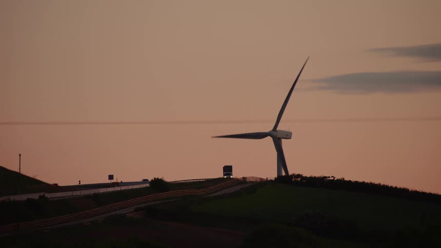 Wind turbine next to a higher area of land with a road where cars are passing outdoors during dusk or dawn