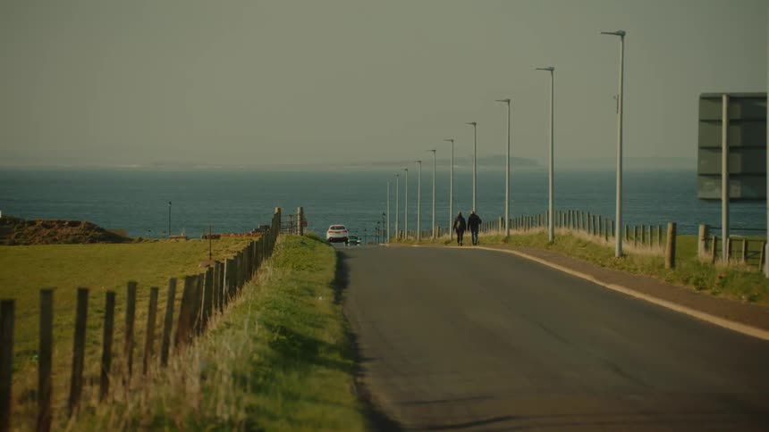 Two adult people walking next to a road where cars are passing close to the ocean outdoors during day time