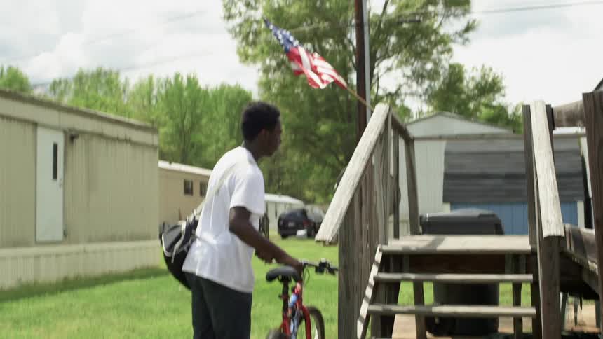 A boy rides a bicycle up to a mobile home and approaches the front door