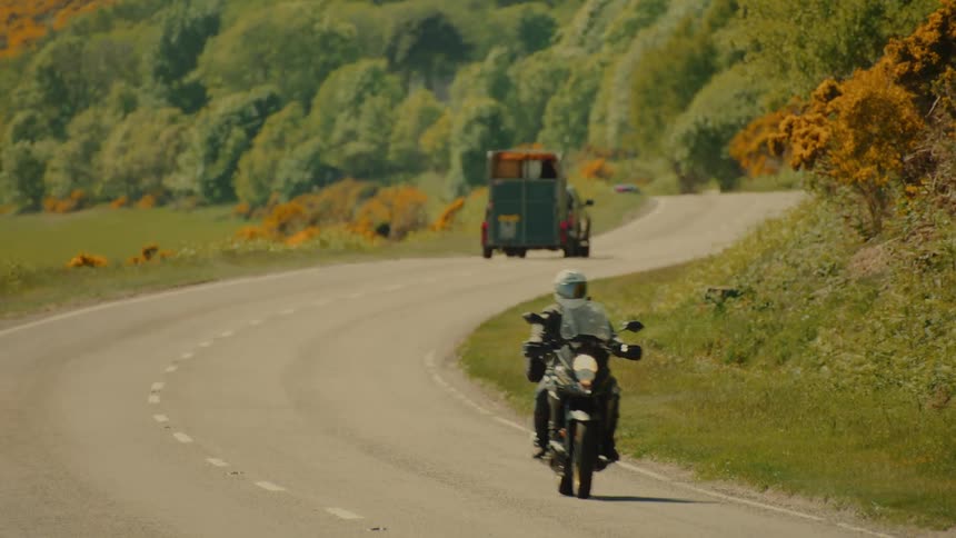 Motorcyclists ride along a winding road through a rural area with dense vegetation