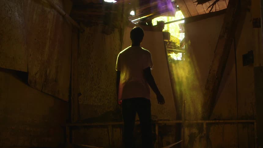 A boy stands inside an abandoned building with sunlight streaming through the ceiling