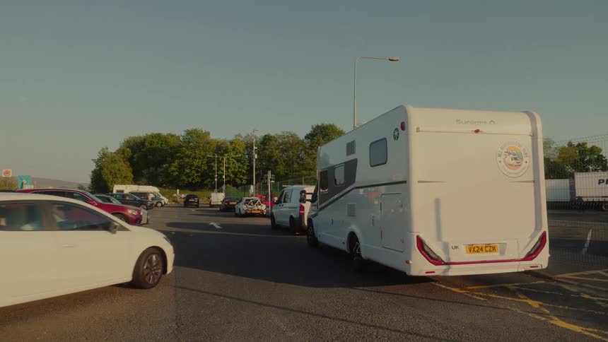 A white motorhome with a united kingdom sticker parked in a large asphalt lot with several other cars and vans under a clear sky during the day