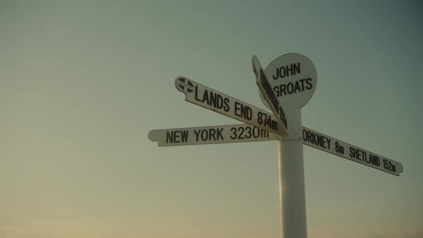 A white signpost with multiple directional arrows pointing to famous locations and their distances like lands end new york orkney and shetland stands against a clear soft hued sky