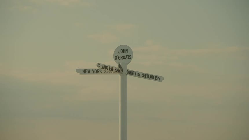 A white signpost with multiple directional arrows stands on a gravel surface against a pale sky during the day