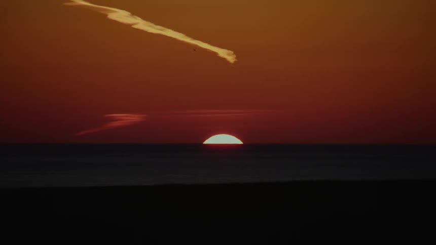 The sun begins to set over the dark ocean horizon creating a deep orange and red sky with a single long cloud stretching across the top during a calm evening