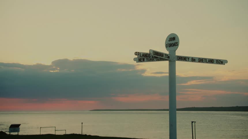 A road sign on the coastline pointing in several directions with labeled signs and a lake view in the background on a cloudy sunset evening