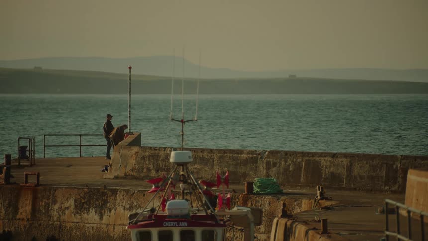 A red and white boat sits in a harbor near a stone pier where two people stand looking out at the calm blue ocean toward rolling hills under a hazy sky during the late afternoon
