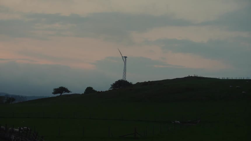 A lone wind turbine stands on a dark green hill under a cloudy and grey sky during a calm dusk
