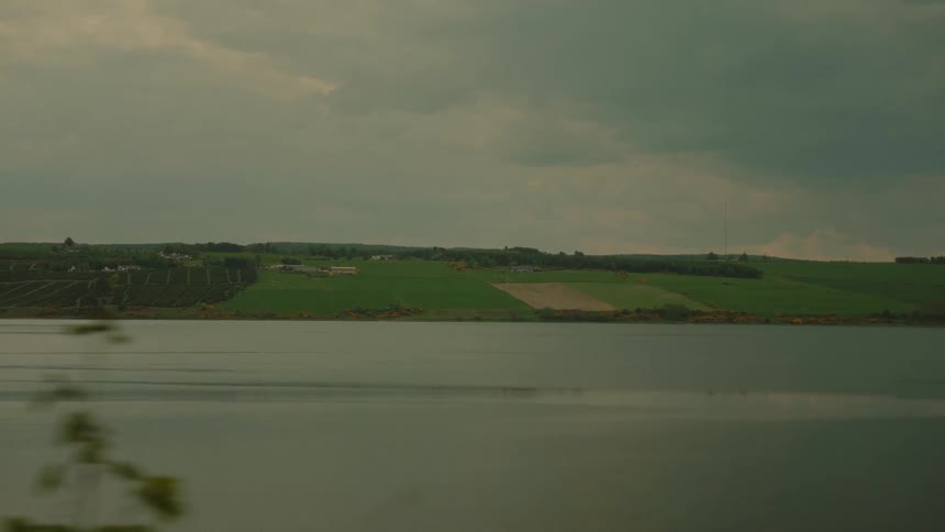 Lush green hills and a calm lake pass by the window of a moving vehicle