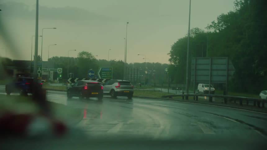 A line of cars navigates a rainslicked roundabout during a gloomy overcast dayrhythmically moving windshield wipers frame the view of the wet asphalt and distant road signs