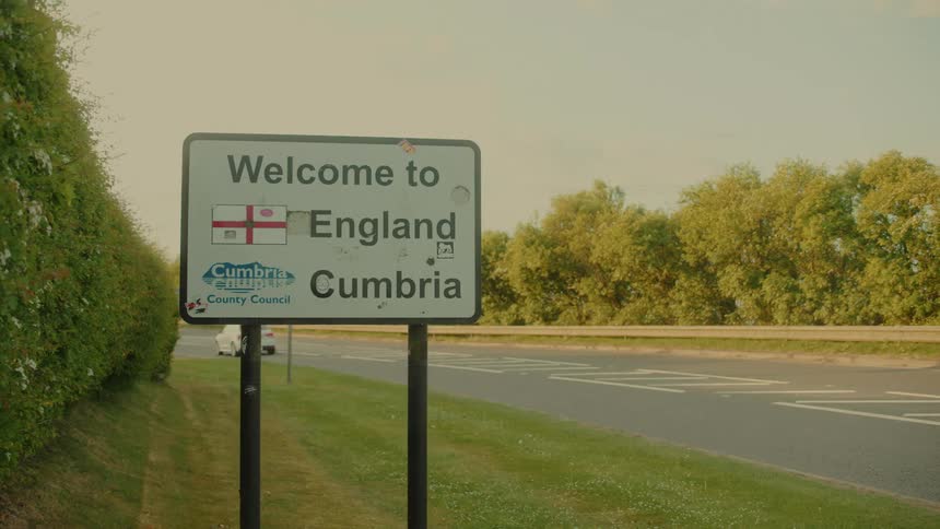 A road sign at the english border with vehicles moving on the road in the evening