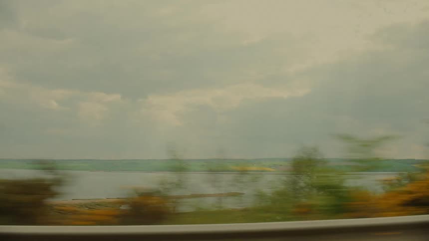 A view through a vehicle window of green fields and a body of water passing by under a dark and cloudy sky during a storm