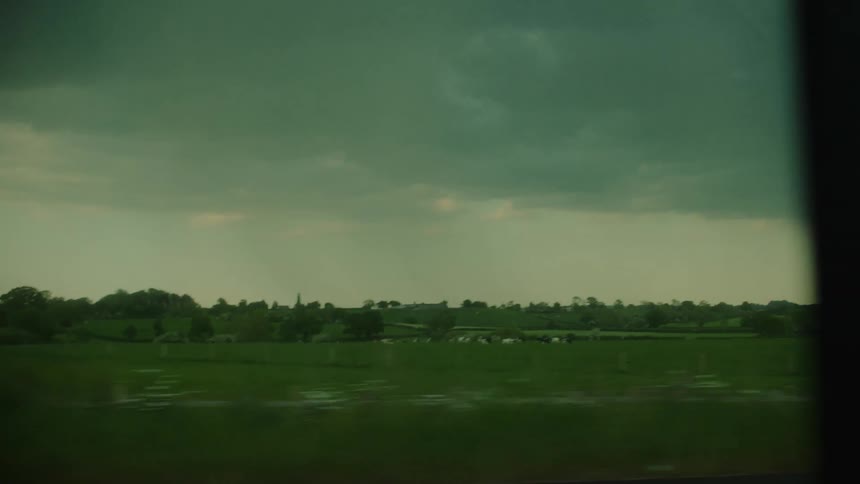 A view through a vehicle window of green fields and trees passing by under a dark and cloudy sky during a storm