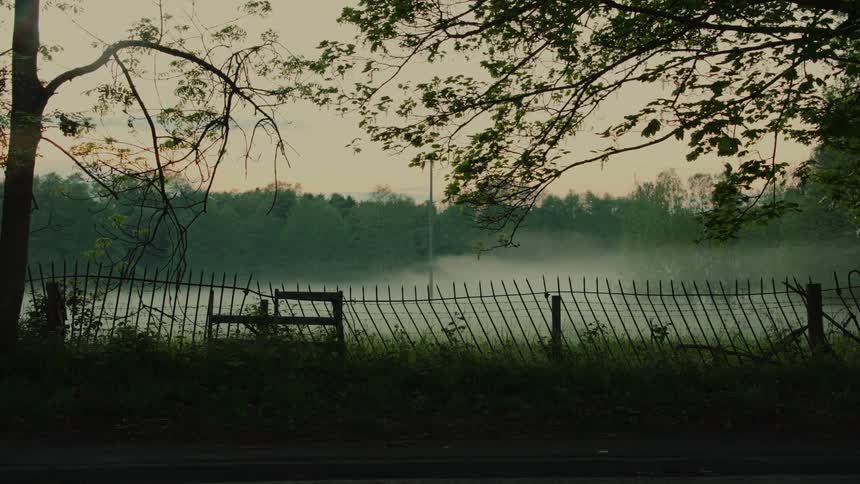 A thick layer of morning mist hangs over a still lake and green field behind a metal fence