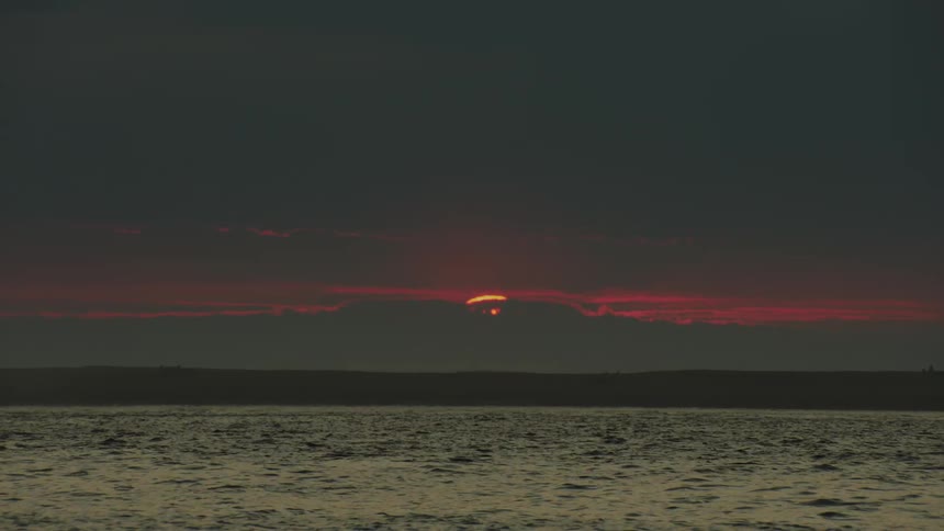 Seagulls fly over the water along the coast as the sun sets over mountains in the distance