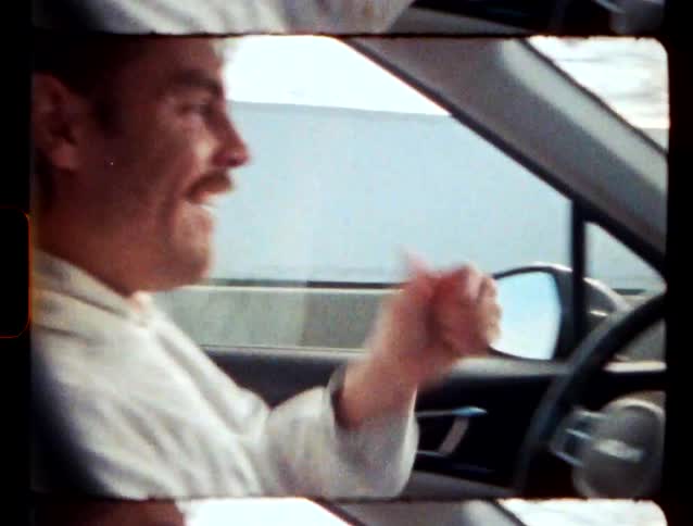 Young adult man wearing a hoodie driving a car while smiling and giving a thumbs up during day time