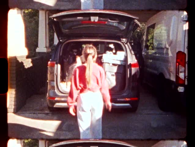 A young girl in a red sweater waves toward a black vehicle with an open trunk before it closes in a driveway during the day