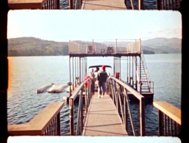 A person walks along a wooden pier toward a boat on a calm lake surrounded by mountains under a clear sky during the day