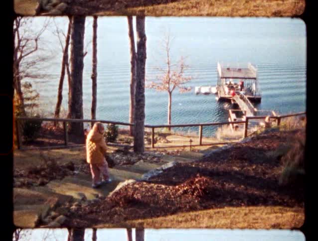 Child with long blonde hair wearing a coat walking down a slope next to the river towards a pier where people are sitting down outdoors during day time