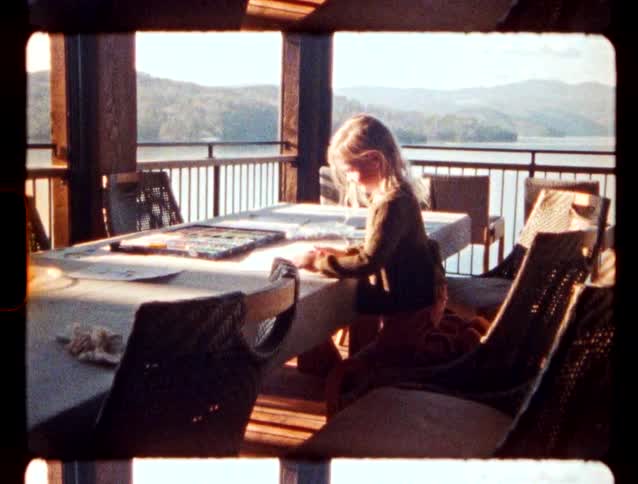 Girl kneeling on a chair drawing on a table on a porch with a lake in the background