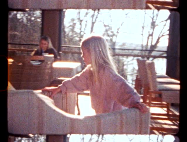 Blonde girl placing her hands on an upholstered chair on a porch with tables and a lake in the background