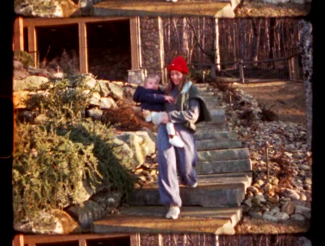 A woman wearing a red beanie and grey loungewear carries a small child down a set of stone steps outside a house the footage has a vintage filmlike aesthetic with visible sprocket holes and a warm grainy texture