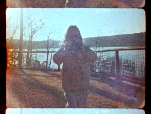 Girl taking a photograph in front of a lake with metal railings and hills in the background