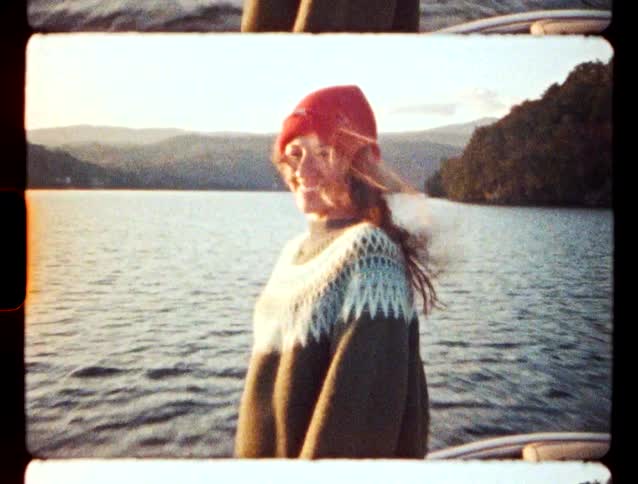 A smiling woman wearing a red beanie and patterned sweater stands on a boat with a lake and mountains in the background