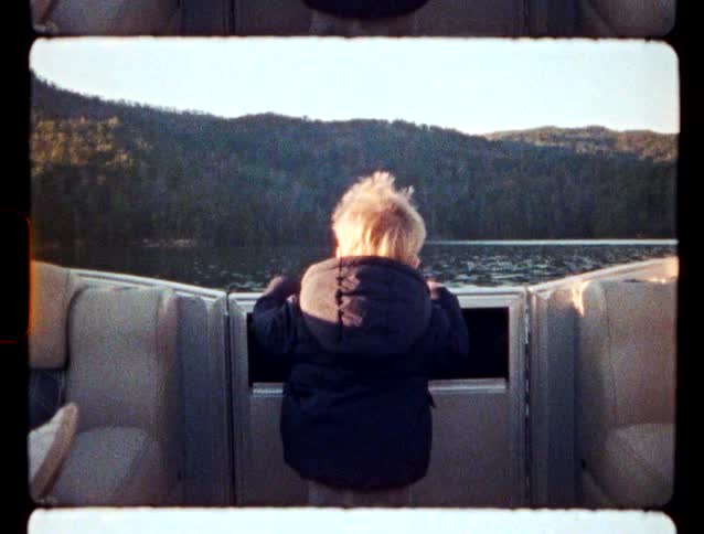A young child in a dark jacket stands on a boat and looks out at the water and distant mountains during the day