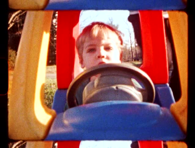 A young boy sits behind the steering wheel of a colorful plastic toy car in a sunny backyard