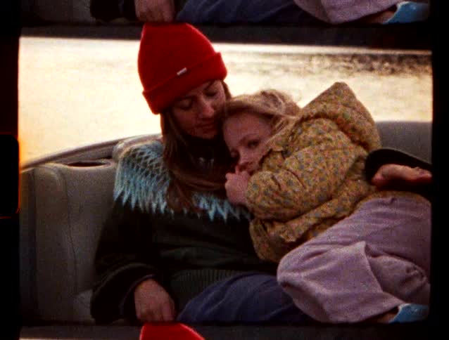 Mother and daughter cuddle while riding in a vehicle on the water under a red tinted sky during winter