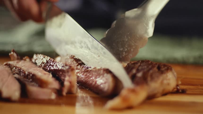 A man uses a sharp knife and metal tongs to slice a piece of grilled medium rare steak into thin strips on a wooden cutting board in a kitchen setting