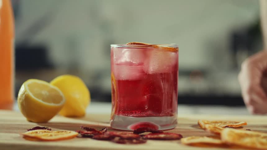 A hand reaches into frame to place a dried orange slice onto the rim of a red iced drink then picks up the glass from the wooden board