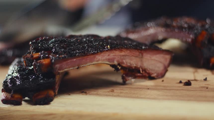 A person is blurred in the background using tongs on ribs ribs are sitting in the foreground