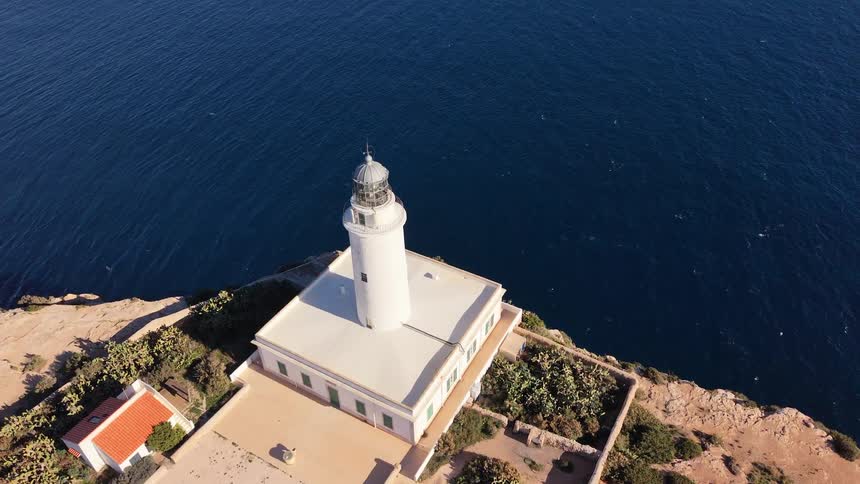 An aerial view shows a white lighthouse and nearby small houses on a rocky coastline overlooking a vast blue ocean