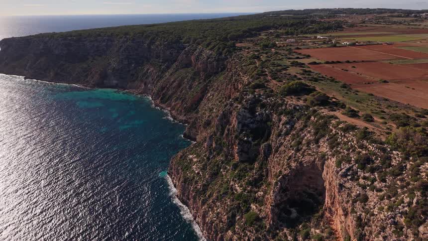 An aerial camera glides along a massive limestone cliff where the deep blue sea meets a rugged coastline
