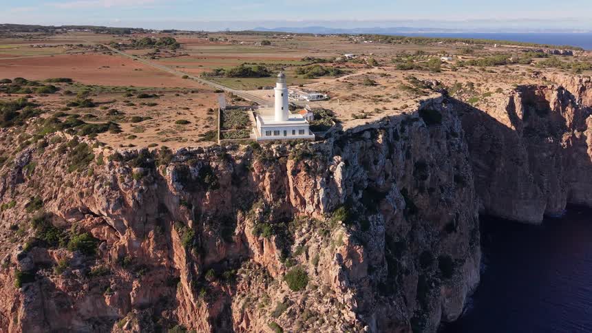 A white lighthouse sits on the edge of a high cliff overlooking a blue ocean under a clear sky during the day