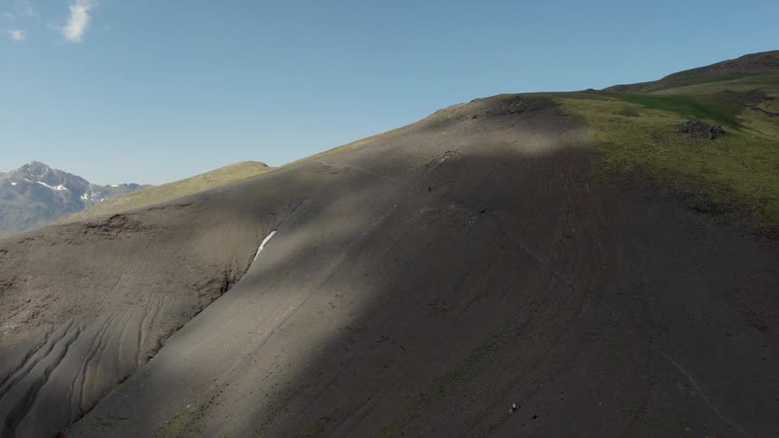 An aerial drone shot captures a wide expanse of a steep mountain slope covered in dark volcanic earth and loose scree under a clear blue sky