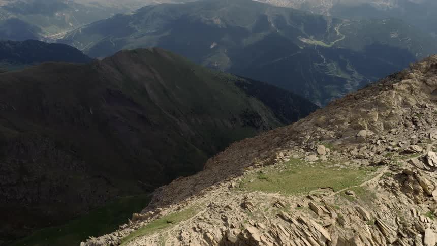 This aerial footage captures a rugged rocky mountain ridge in the foreground with a vast valley belowthe camera pans to reveal a series of distant hazy peaks stretching toward a cloudy horizon