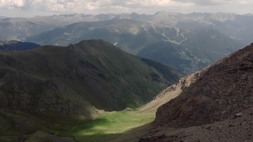 This video features a sweeping wide shot of a vast mountain range under a cloudy sky the camera slowly pans across the rugged rocky peaks and a lush green valley nestled between the slopes